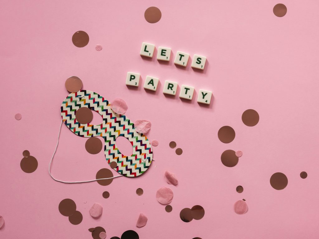 A colorful party mask with confetti and Scrabble tiles spelling "LET'S PARTY" on a pink background.